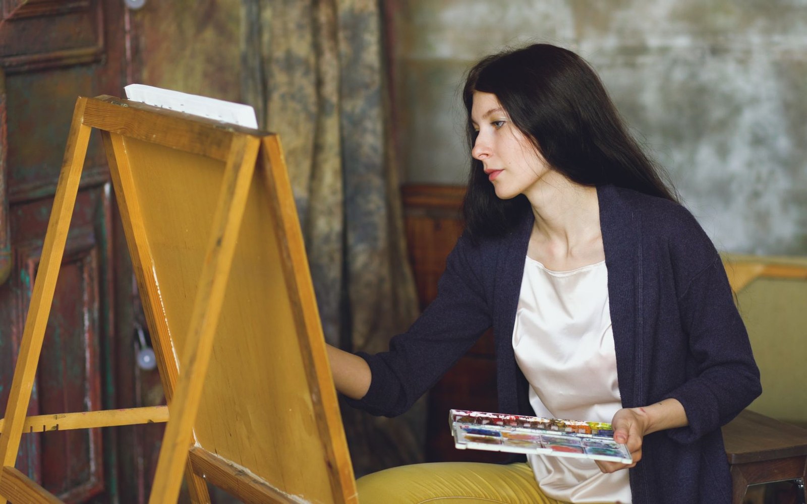 Adult painter at a large canvas in a north-lit studio, palette in hand, adult model visible on the stand behind them