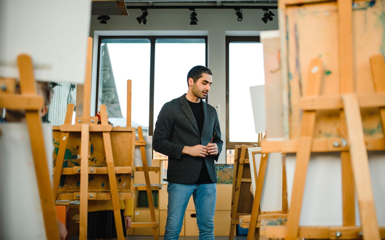 A man in a blazer stands among wooden easels in a bright art studio