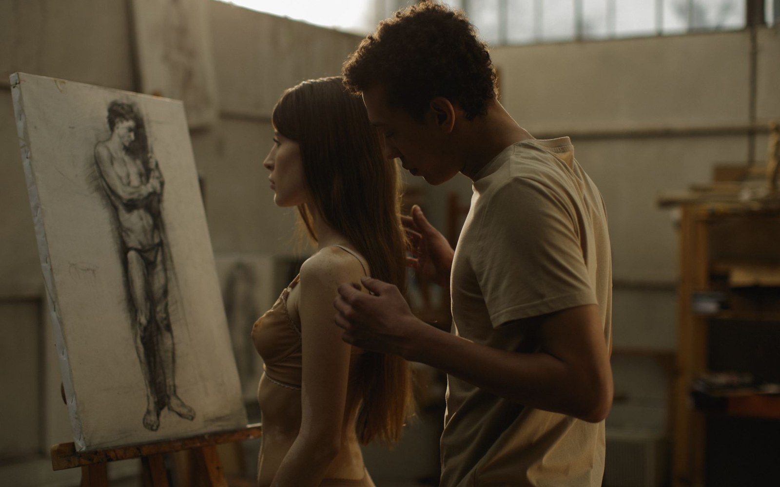 A man rests his forehead against a woman's head beside an easel with a figure sketch