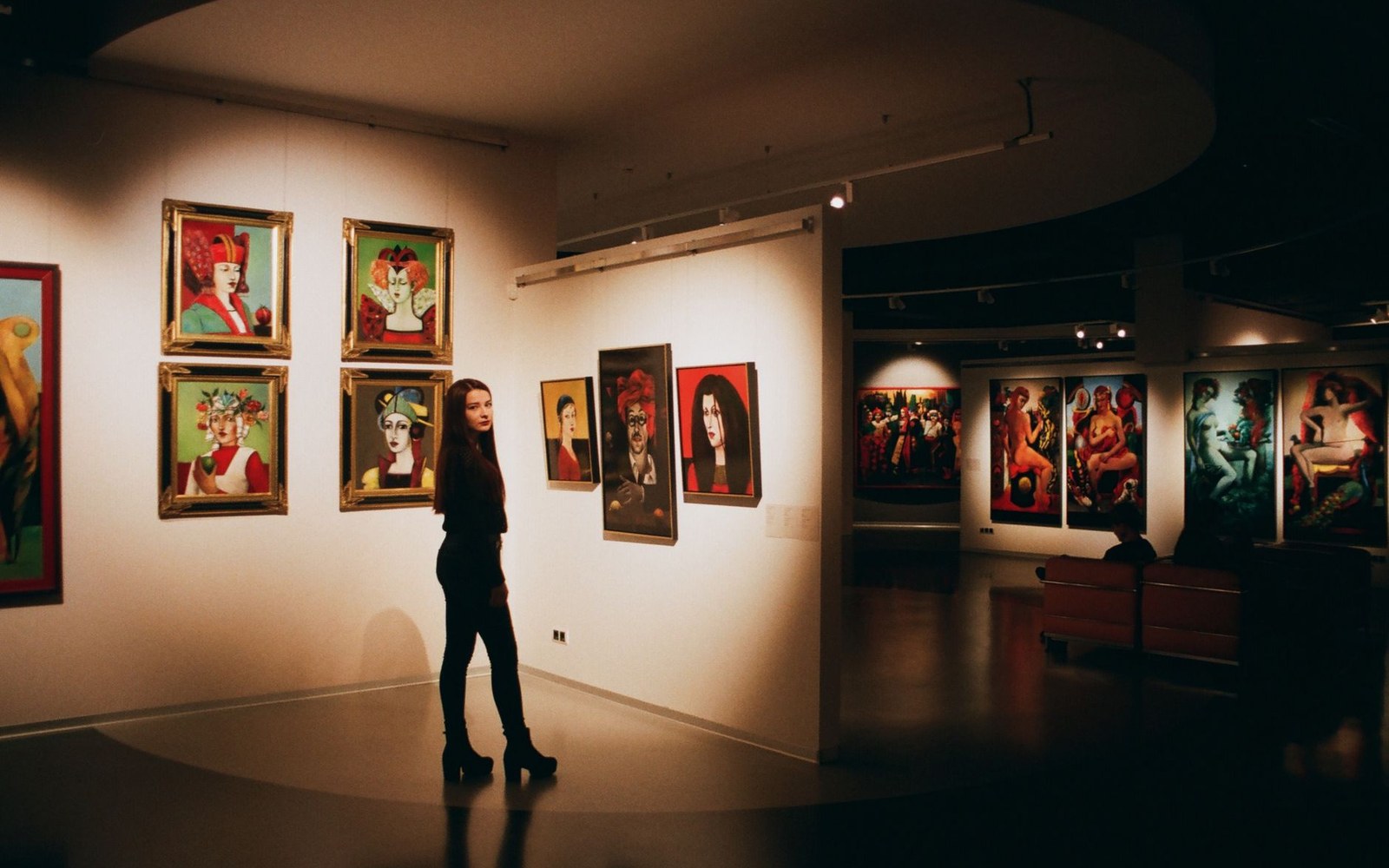 Woman stands viewing colorful portrait paintings in a dimly lit gallery