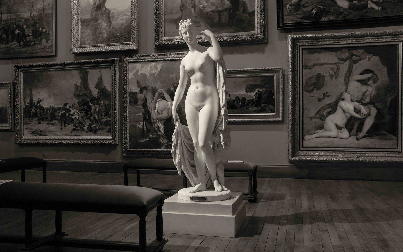 A marble statue of a nude woman stands on a pedestal in a gallery lined with framed paintings