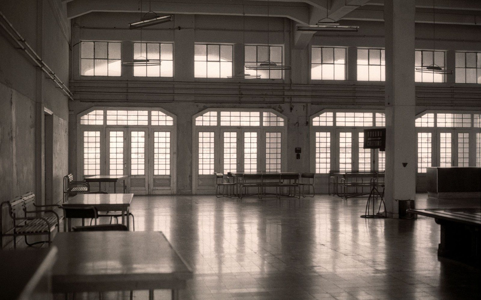Empty industrial hall with tall arched windows and scattered tables and chairs